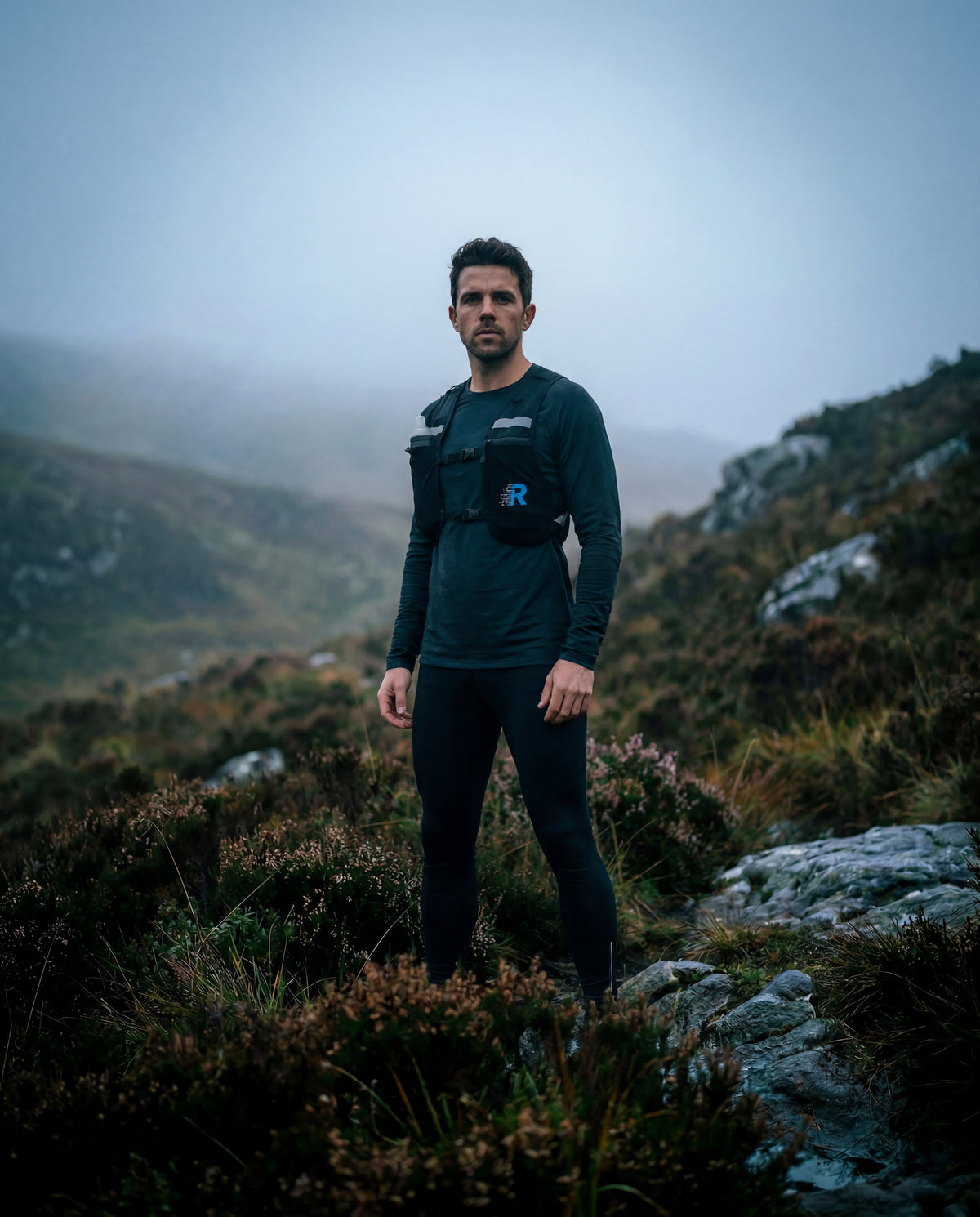 Man standing in a mountainous landscape wearing a dark outfit with a logo - - wearing the run with freedom challenger running vest with hydration flask
