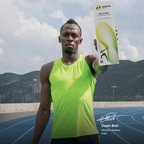 Usain Bolt stands on an outdoor running track holding a box of Exertor Walking shoes, with mountains and a cloudy sky in the background.